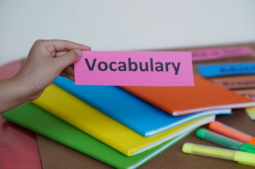 Hand hält ein Schild mit der Aufschrift „Vocabulary“; im Hintergrund sind verschieden bunte Hefte uns Stifte